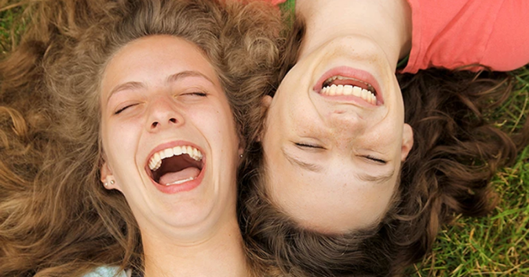 Two young women laughing on grass, eyes closed.