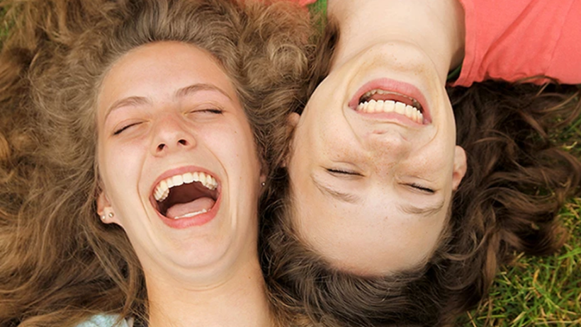 Two young women laughing on grass, eyes closed.